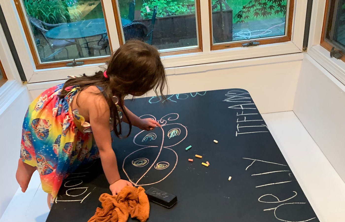 A young girl in a colorful dress draws with chalk on a black table