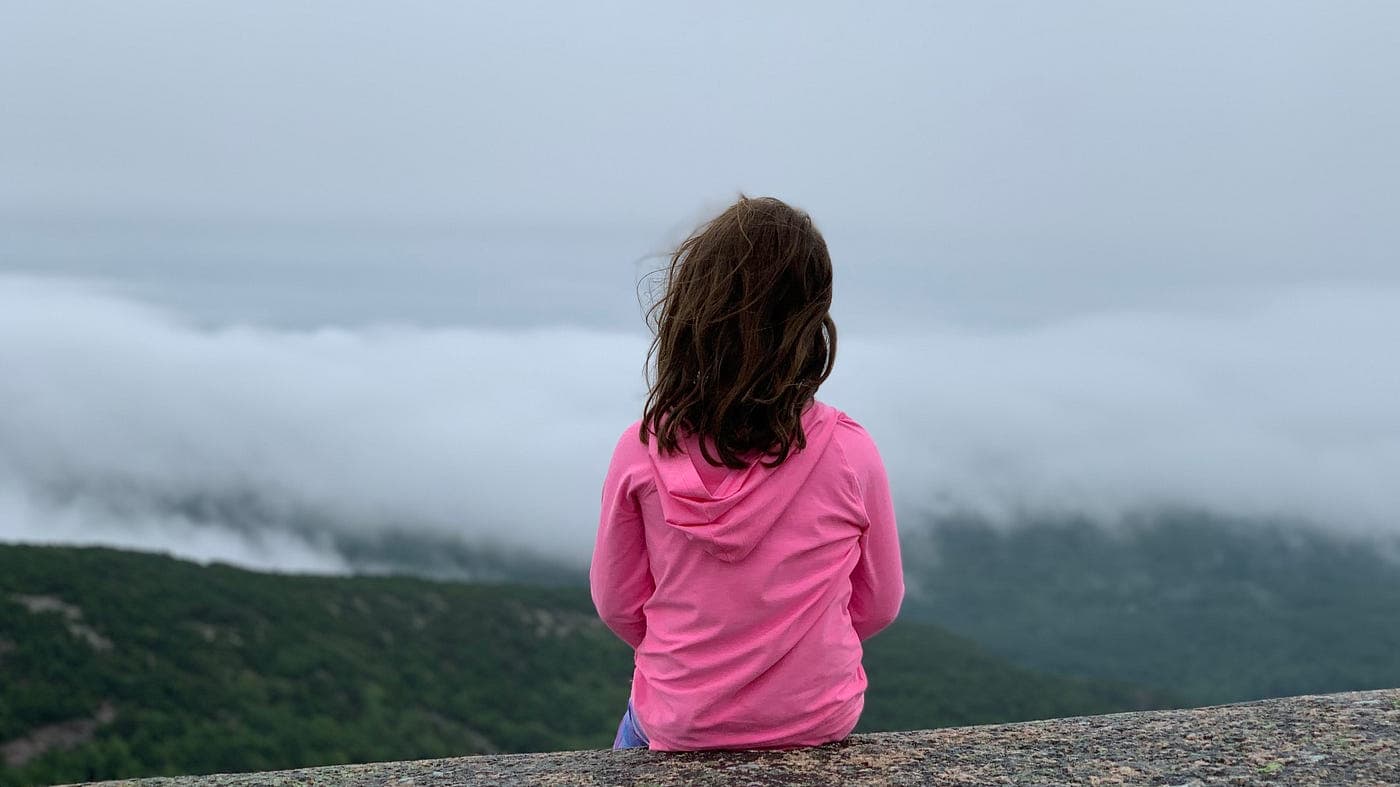 A young girl in a bright pink hoodie looks out over a sea of fog atop rolling hills